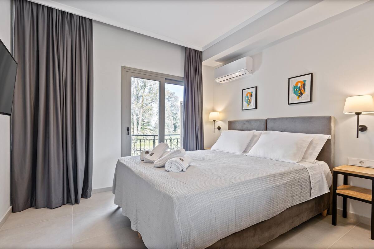 A modern hotel room with a large bed, a bedside table, a lamp, and a view of the outdoors.