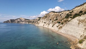 A coastal landscape with clear blue waters, sandy beach, and rocky cliffs. - Comissa Luxury Suites (Corfu)