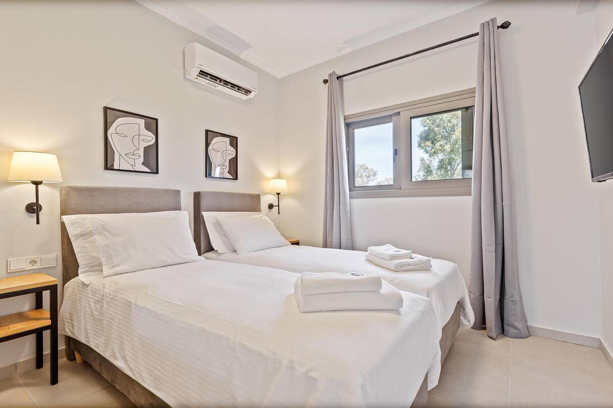 A hotel room with two beds, a window with curtains, a wall-mounted air conditioner, and two framed artworks.