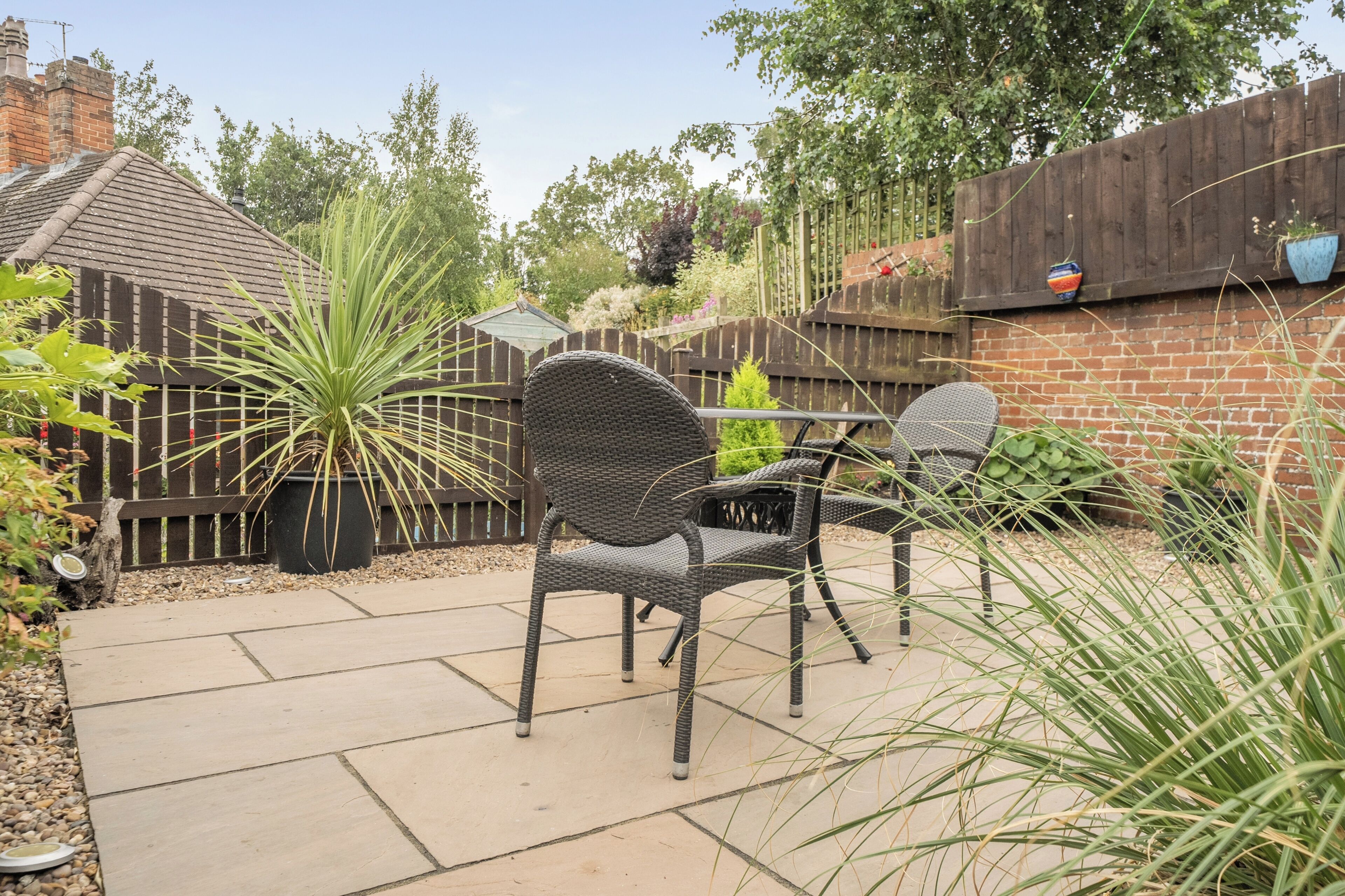 Een terras met een rieten stoel en een tafel, omgeven door een houten hek en diverse planten.