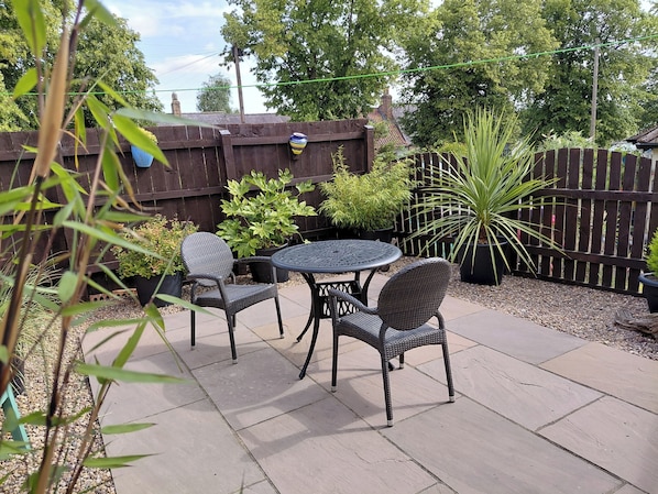A patio with a round table and two wicker chairs, surrounded by potted plants and a wooden fence.