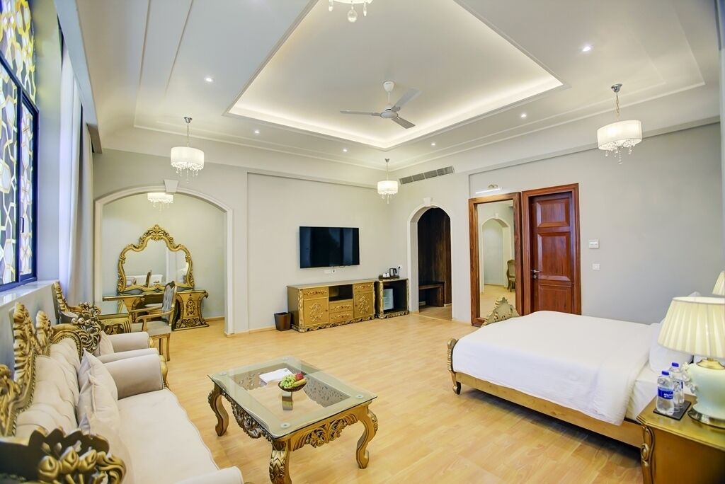 A spacious hotel room with a large bed, a sitting area with a sofa and a coffee table, a television, and a chandelier.