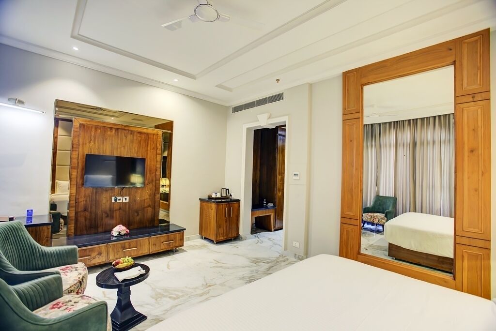 Une chambre d’hôtel moderne avec un grand lit, un téléviseur fixé à un meuble en bois, une petite table avec des fruits et un miroir agrémenté de rideaux.