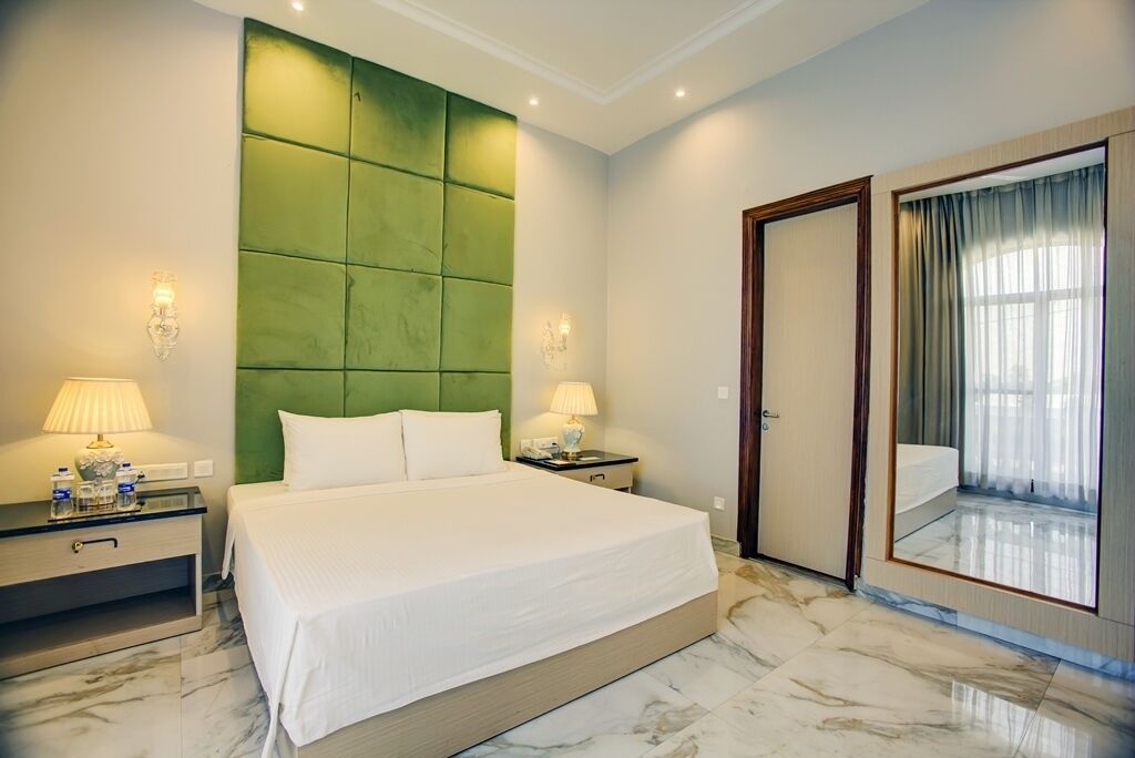 A hotel room with a large bed, two bedside tables with lamps, a green padded headboard, a large mirror, and a door leading to a balcony.