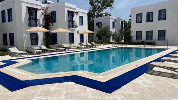 A swimming pool with lounge chairs and umbrellas, surrounded by white buildings with blue trim. - Ege Apartments (Bodrum)