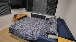 A modern bedroom with a large bed, a TV mounted on the wall, and a window with blinds.