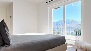 Un dormitorio con cama, un banco blanco mullido y un amplio ventanal corredera que da a un balcón con vistas a la montaña.