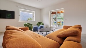 Una sala moderna con un amplio sofá modular, una mesa redonda de centro y un televisor montado en la pared. Hay dos ventanas que ofrecen luz natural y vistas al exterior.