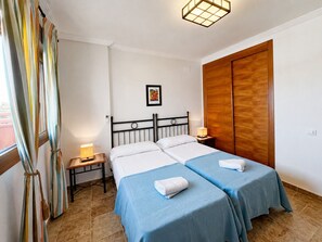 A bedroom with two beds, a wooden wardrobe, a bedside table with a lamp, and a window with curtains.