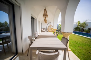 Uma área de refeições com uma mesa comprida e cadeiras, uma cadeira suspensa, e vista para uma piscina.