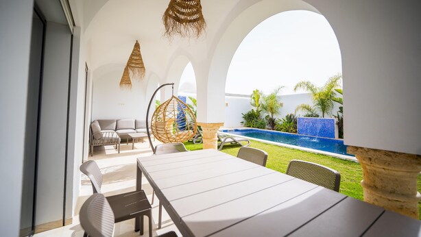 Un área de comedor al aire libre moderna con vista a la piscina, que incluye una mesa larga, sillas e lámparas colgantes de ratán.
