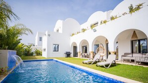 Un área de piscina moderna con una piscina azul cristalina, arquitectura blanca y mobiliario exterior.