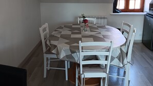 Zona pranzo con tavolo rotondo, sedie bianche e tovaglia fantasia. Si vede una finestra con vista sull'esterno, un termosifone e un angolo cottura con frigorifero.