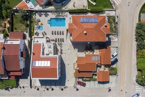 Vue aérienne d’un quartier résidentiel avec une piscine, plusieurs bâtiments aux toits recouverts de tuiles rouges et une route.