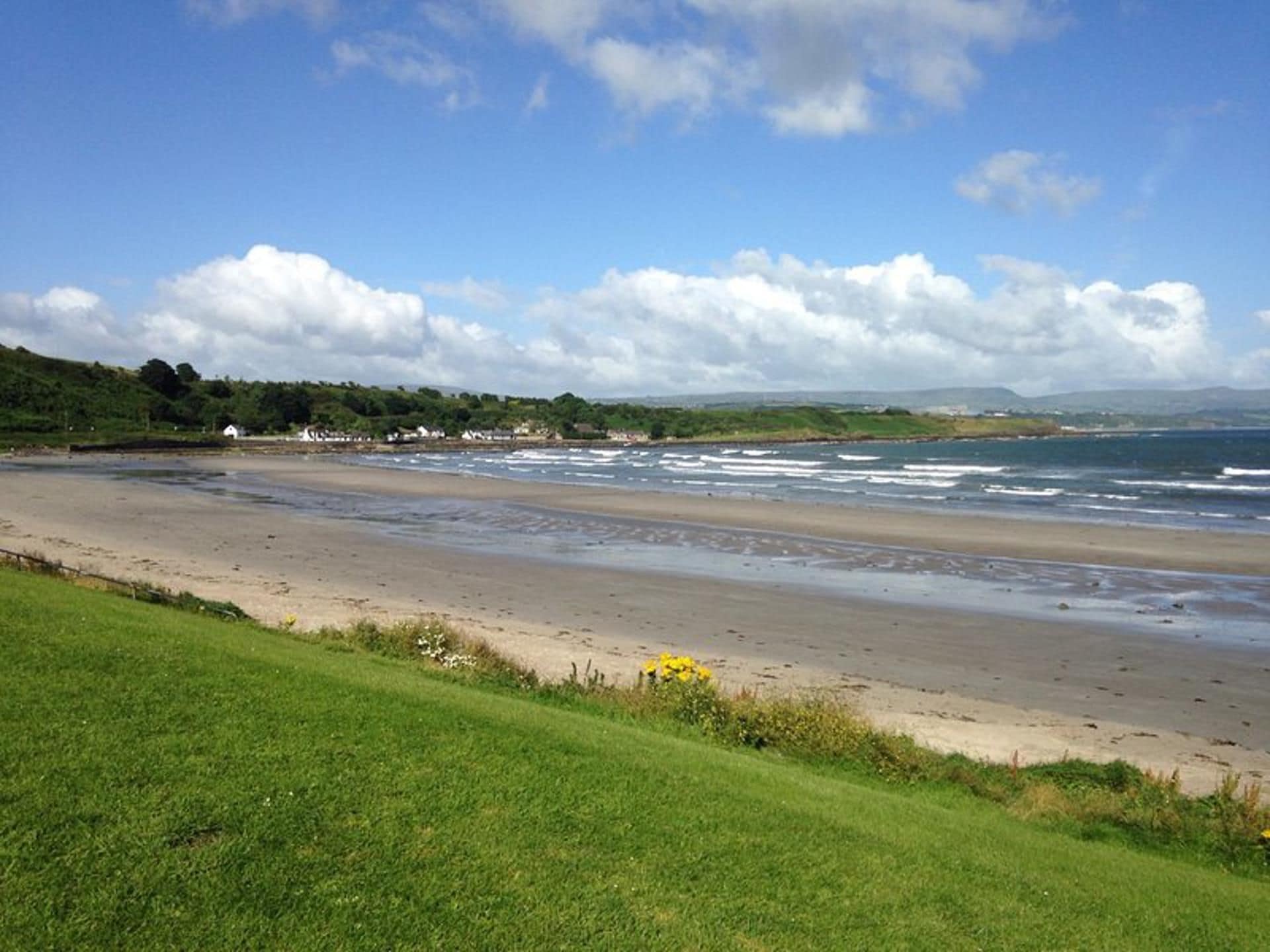 A coastal landscape with a sandy beach, grassy areas, and a distant view of hills under a partly cloudy sky.