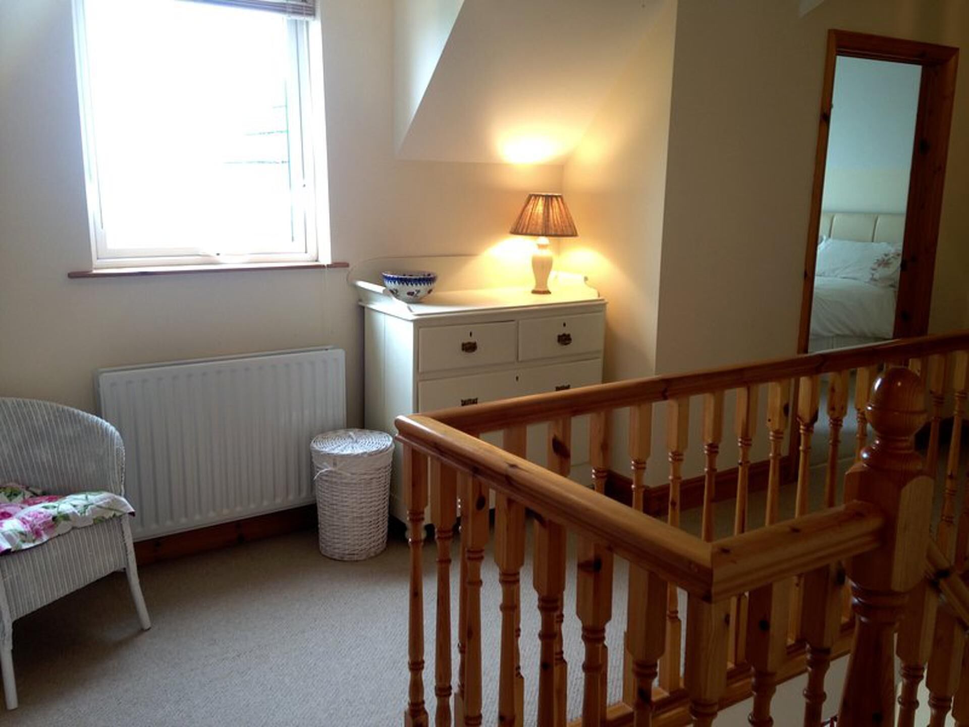 Une chambre à coucher comprenant un lit, une commode, une lampe, une chaise, un radiateur, une corbeille à déchets et une rambarde d’escalier en bois.