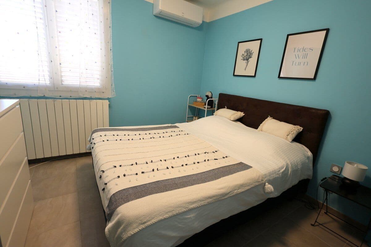 A bedroom with a bed, a radiator, a window with blinds, and two framed pictures on the wall.