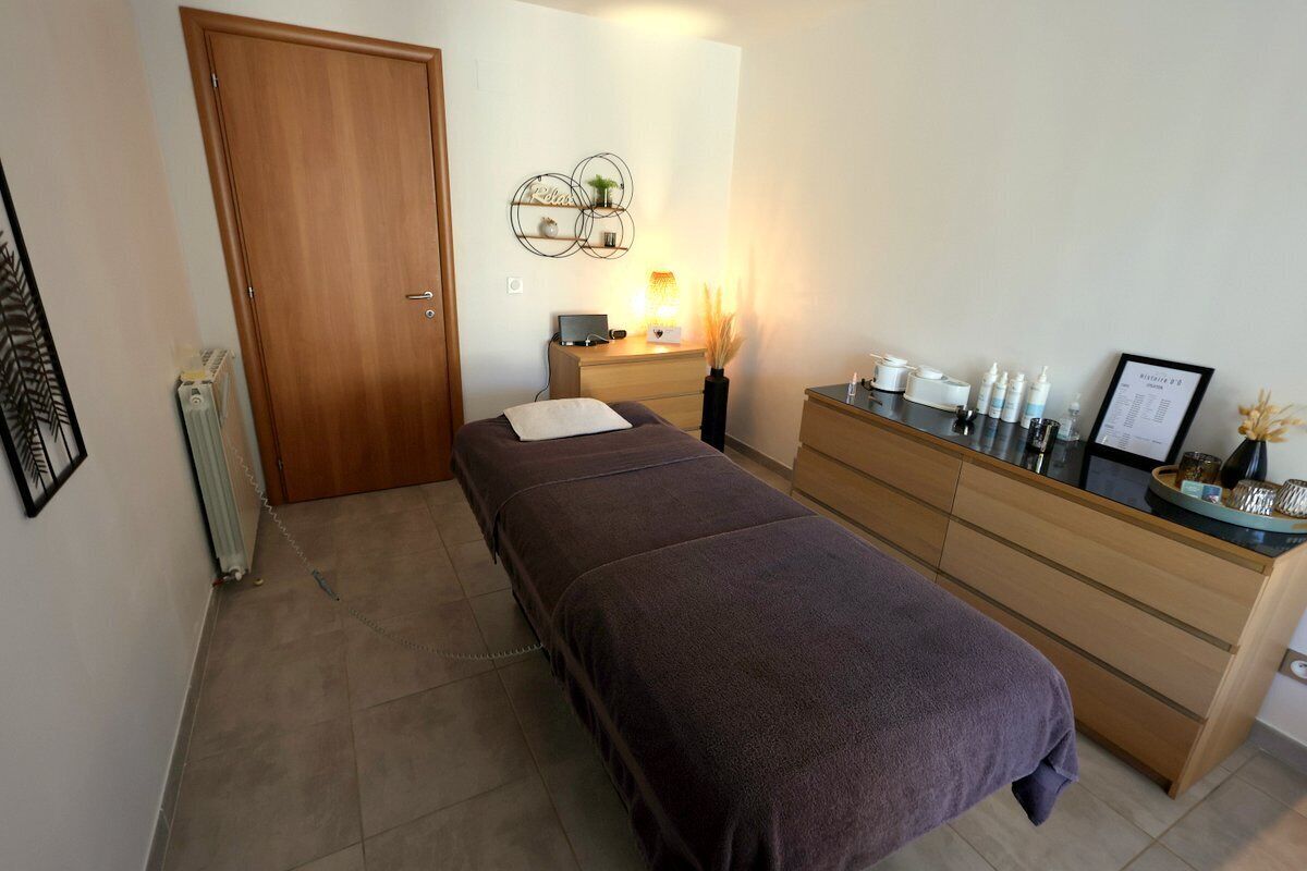 A bedroom with a bed, a dresser, a desk with a lamp, and a wall-mounted shelf with decorative items.