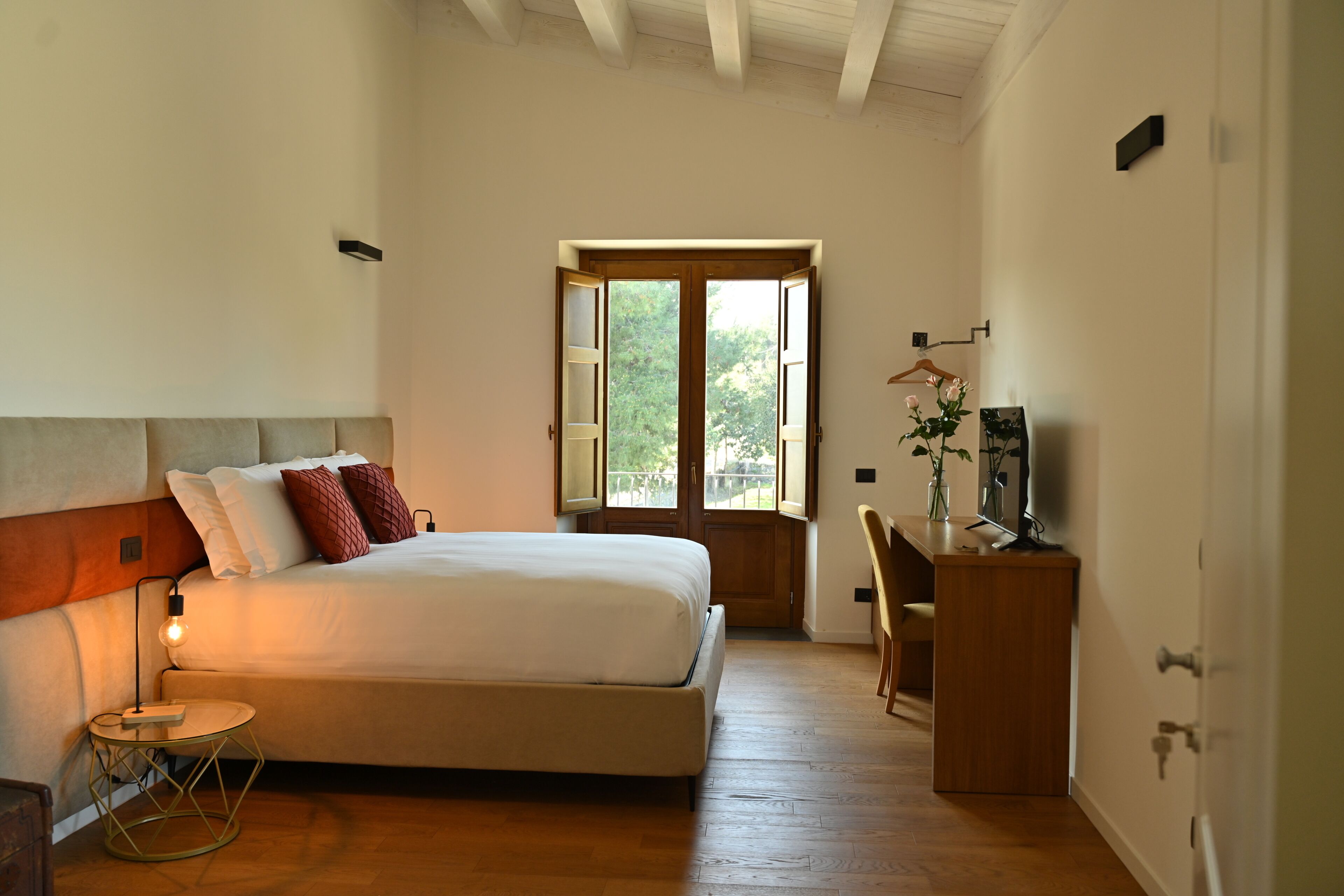 A bedroom with a bed, bedside table, desk, chair, and a window with a view of greenery.