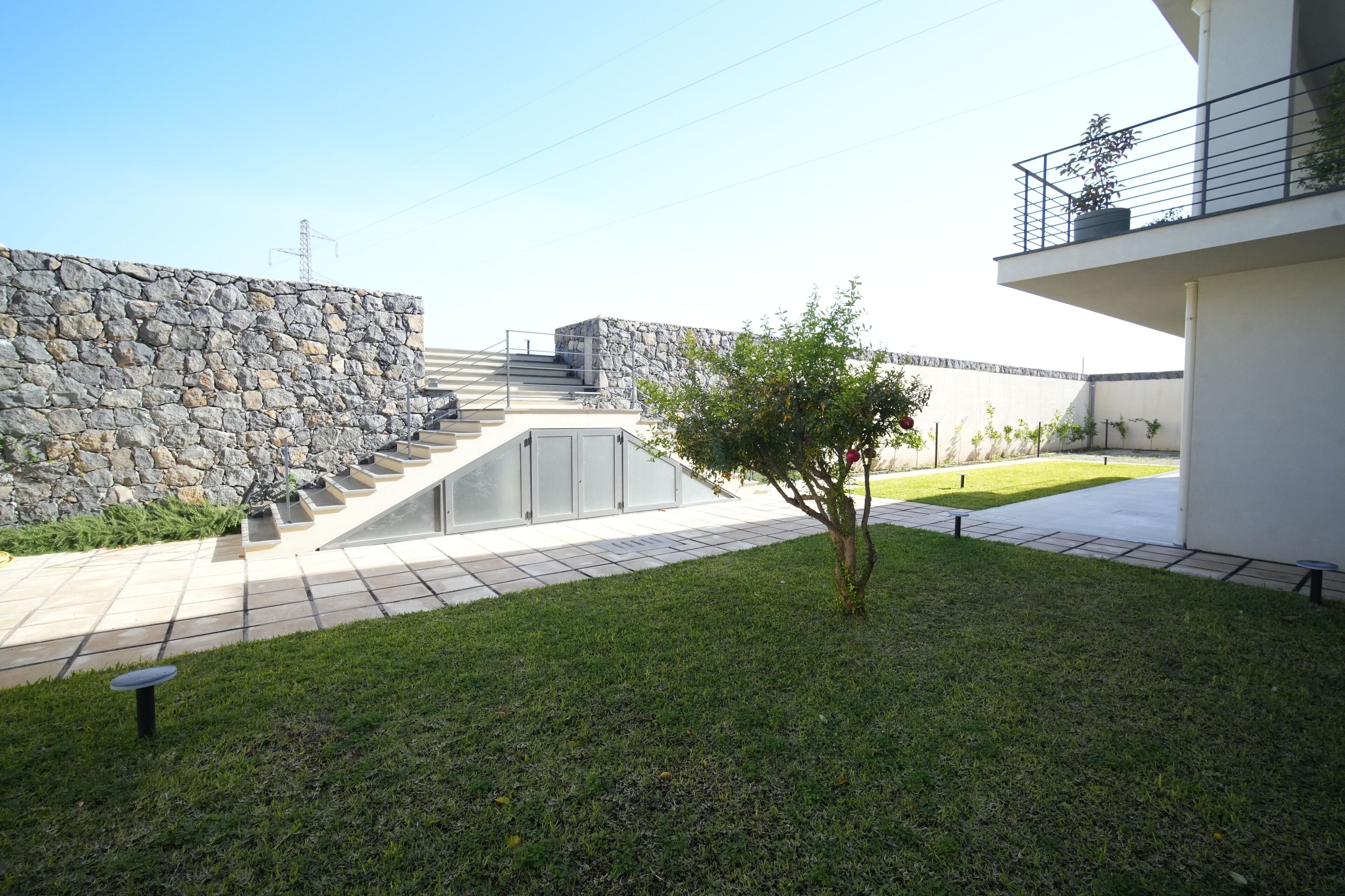 Un edificio moderno con una pared de piedra, un pequeño árbol y un área pavimentada con escalones que conducen a un balcón.