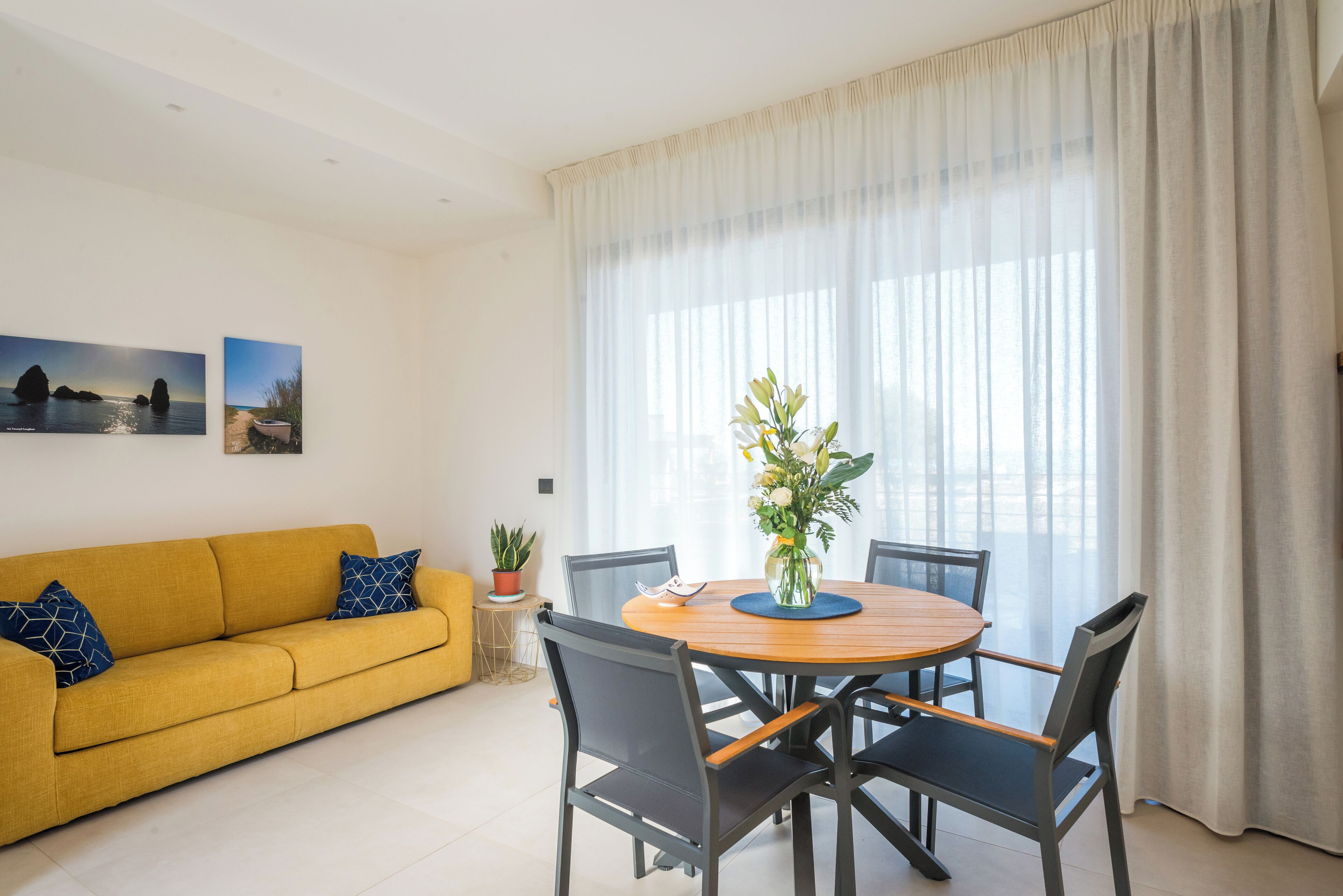 Un salon avec un canapé jaune, une table de salle à manger ronde en bois entourée de chaises, une plante en pot et deux tableaux encadrés accrochés au mur.