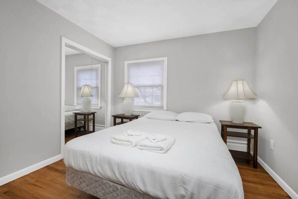 A bedroom with a bed, two bedside tables with lamps, a mirror, and a window with blinds.