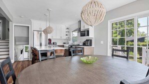 A modern kitchen with white cabinets, a stainless steel refrigerator, a dining table with chairs, and a large window providing a view of the outdoors.