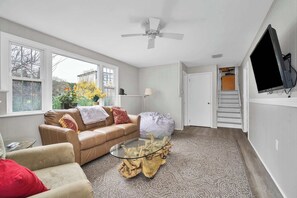 A living room with a glass coffee table, a brown leather sofa, a beige armchair, a large window, a ceiling fan, and a TV mounted on the wall. - Fenwood Coastal Getaway Private Beach Access (Old Saybrook)