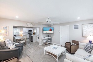A modern living room with a dining area, kitchenette, and a TV displaying a beach scene.