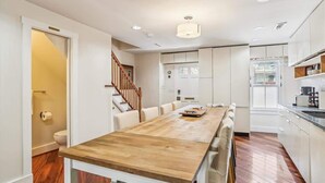 A modern kitchen with white cabinets, a wooden dining table, and a staircase leading to another level. - Downtown Pinkney House w/Parking (Annapolis)