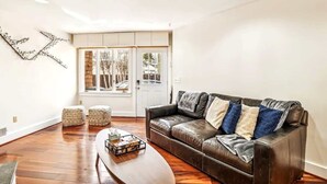 A living room with a brown leather sofa, two ottomans, a wooden coffee table, and a decorative metal wall piece. - Downtown Pinkney House w/Parking (Annapolis)