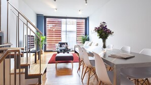 A modern dining area with a long table, white chairs, and a red rug. There is a staircase on the left and a large window with blinds.