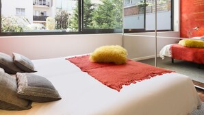 A modern hotel room with a large window, a bed with a red throw, and a view of the cityscape.