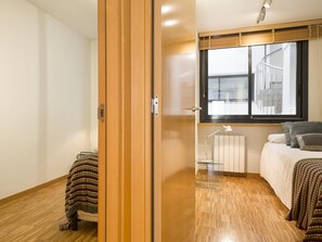 A modern bedroom with a wooden wardrobe, a bed with a patterned blanket, a bedside table with a lamp, and a window with a view of buildings. - Apartment in Sant Gervasi with Balcony for 6 (barcelona)