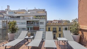 Una terraza en la azotea con varios sillones de sol y plantas en macetas, con vistas a un paisaje urbano de edificios de varios pisos.