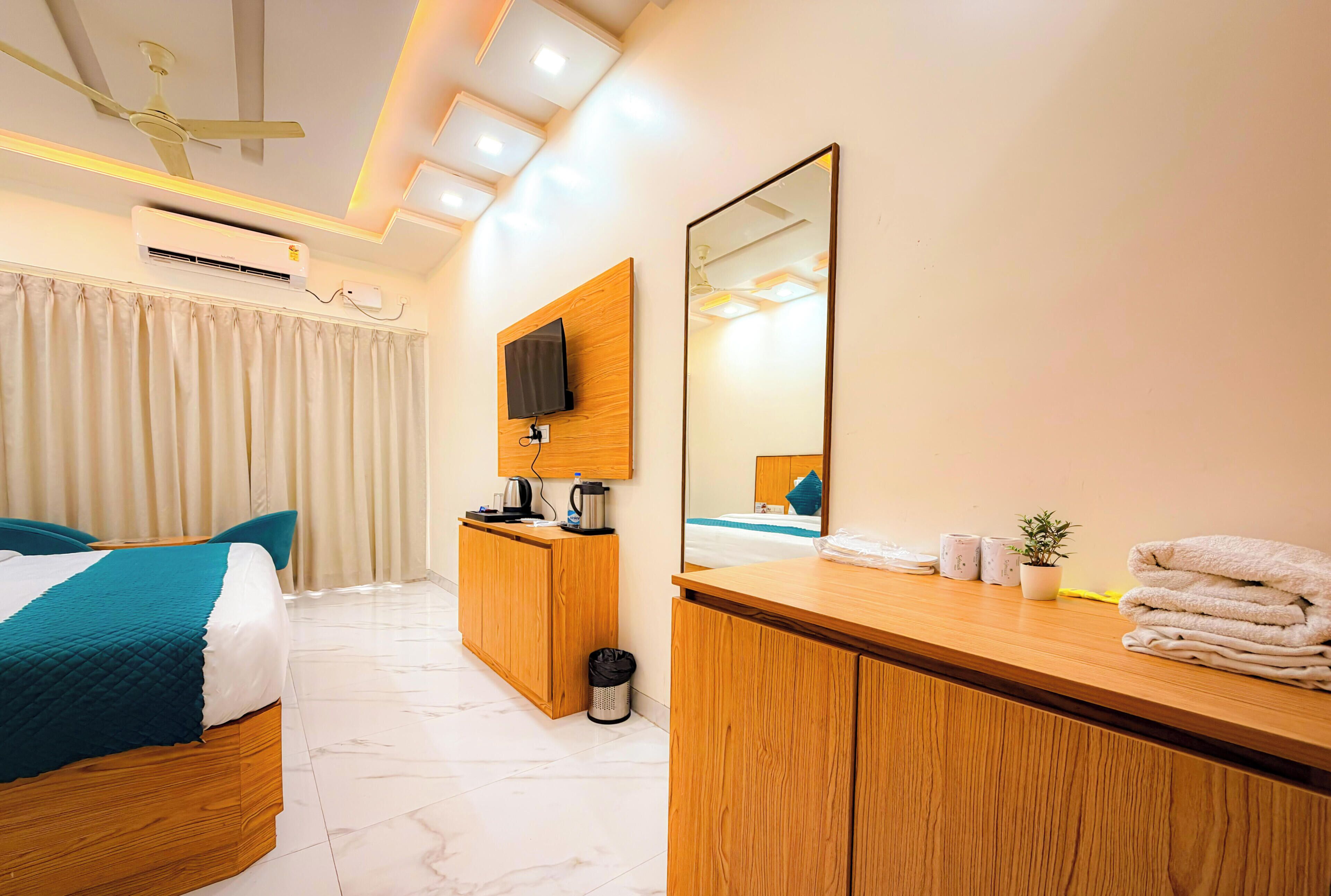 Hotelkamer met een bed, een houten garderobe, een spiegel, een aan de muur bevestigde televisie, een plafondventilator en een kleine plant in een pot.