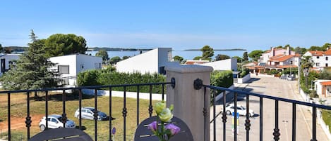 A balcony with a vase of flowers, a view of a coastal town, and parked cars.