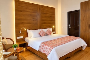 Un dormitorio con cabecera de madera, cama con ropa de cama blanca y una manta floral, una mesita de noche con una planta y una mesita auxiliar.