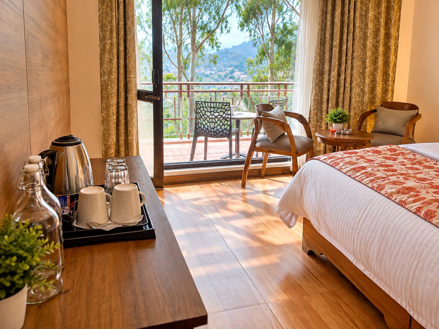 Una habitación de hotel con una cama, una mesa con tetera y tazas, una silla y un balcón con vistas a árboles y colinas.