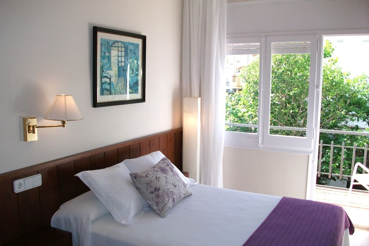 A hotel room with a bed, a nightstand, a wall-mounted lamp, a framed artwork, and a balcony with a view of greenery.