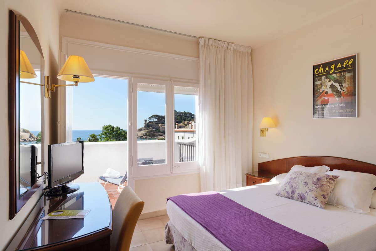 A hotel room with a bed, a desk with a computer, a chair, and a view of the sea.