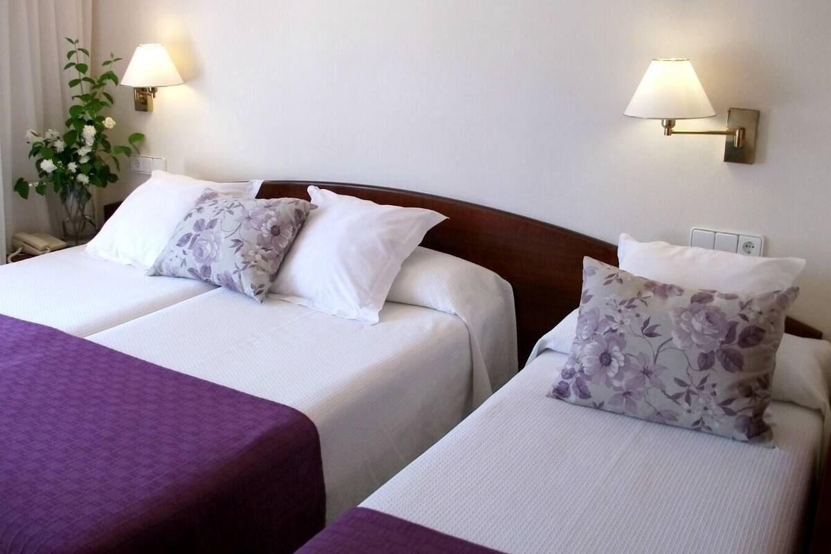 A hotel room with two beds, each with white bedding and floral-patterned pillows. There are wall-mounted lamps on either side of the beds.