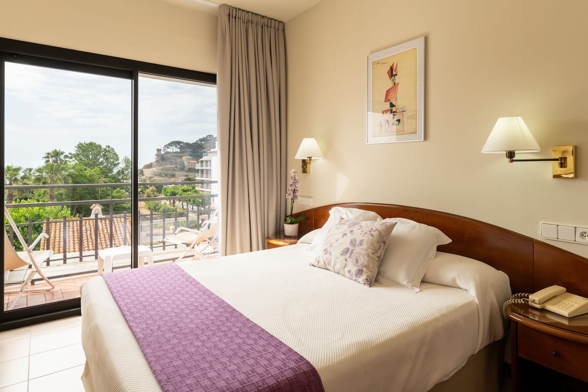 A hotel room with a bed, a balcony, a view of the city, and a painting on the wall.