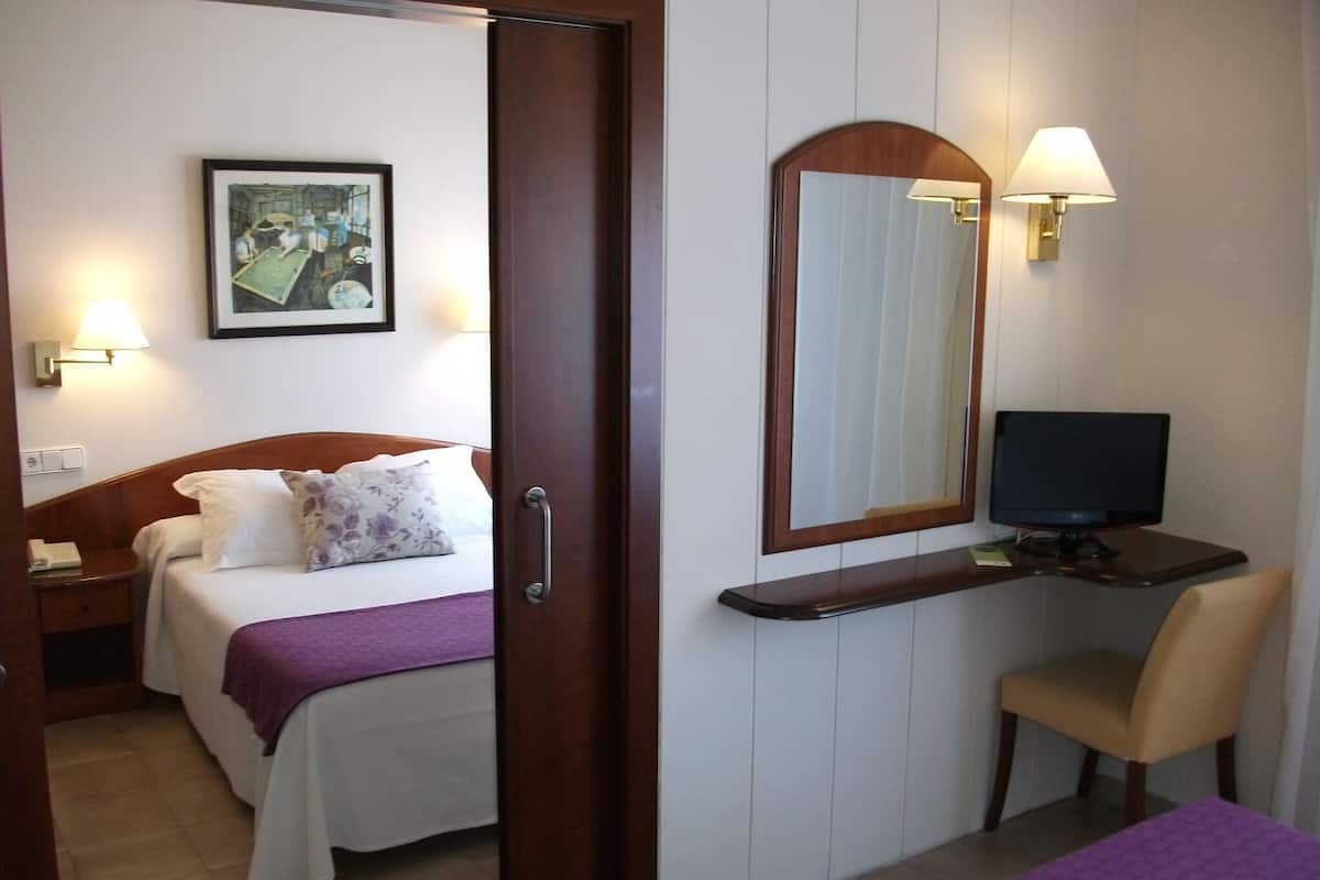 A hotel room with a bed, a desk with a computer, a chair, a bedside table, and a wall-mounted lamp.