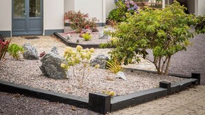 Een woonhuis met twee verdiepingen, een grijs dak en witte muren, een voordeur en een tuin met verschillende planten en rotsen.