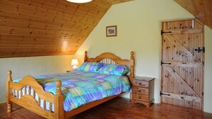 A bedroom with a wooden ceiling, a bed with a colorful quilt, a bedside table with a lamp, and a wooden wardrobe with a sliding door. - Griffins Holiday Cottage (Murreagh)