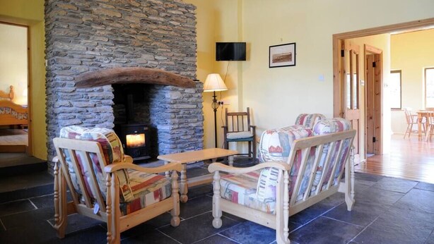 A cozy living room with a stone fireplace, wooden furniture, and a television mounted on the wall. - Griffins Holiday Cottage (Murreagh)