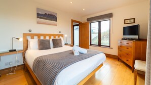 A bedroom with a large bed, wooden headboard, bedside tables with lamps, a dresser, a television, and a window with a view of greenery. - Great Blasket, Dunquin (Dunquin)