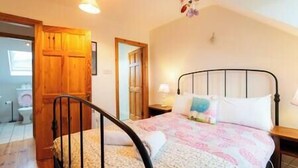 A bedroom with a bed, a nightstand, a wooden door, and a bathroom visible through an open door. - Orchard Cottage - Dingle (Dingle)