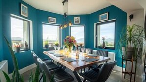 A dining area with a wooden table set for six, surrounded by black chairs. There are potted plants and a vase with flowers on the table.